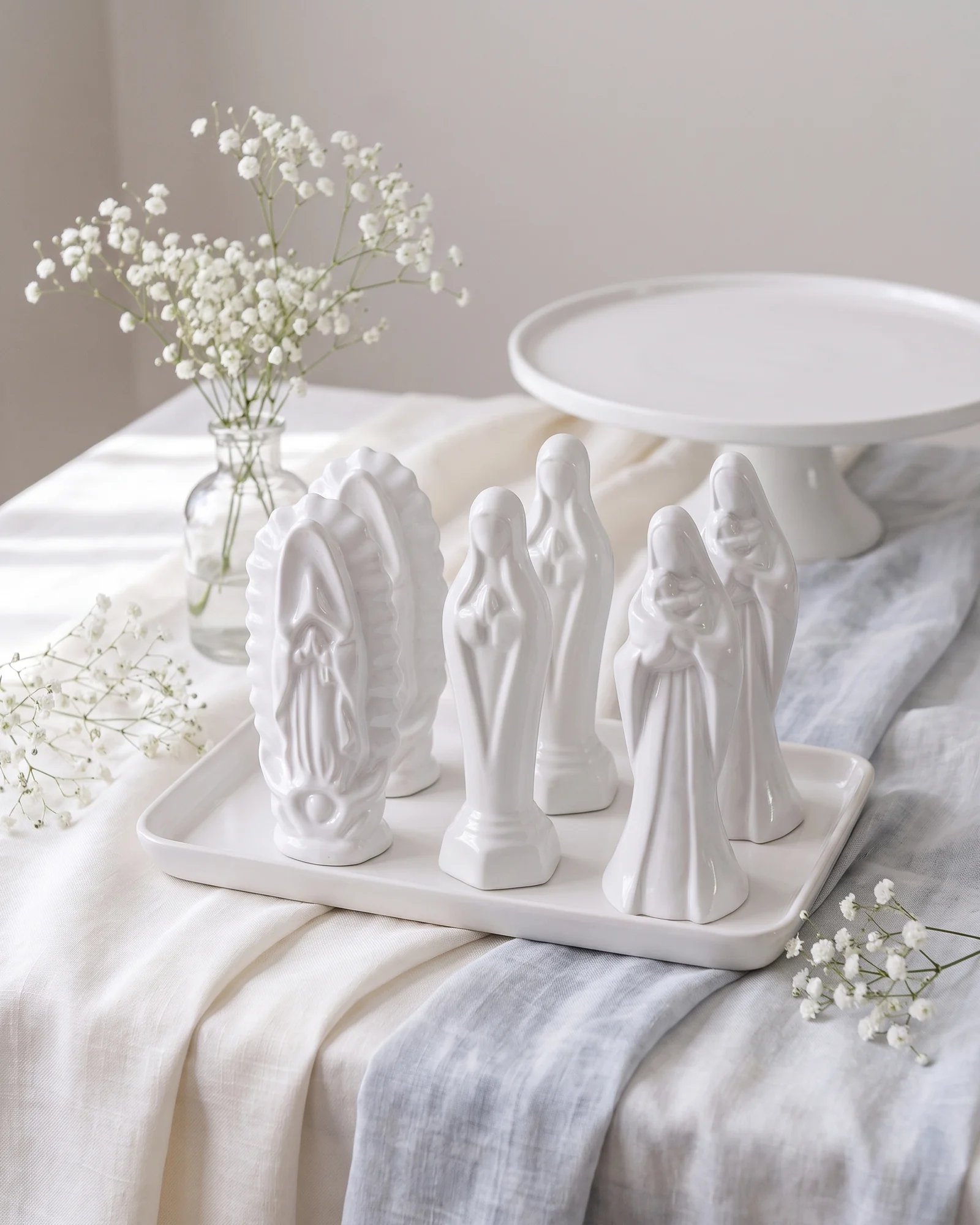 Recuerditos de cerámica blanca de María con el Niño, Madona y Silueta en una charola blanca, sobre una mesa de bautizo con flores nube y lino azul claro.