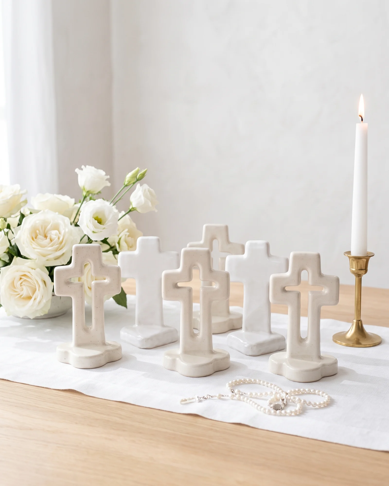 Cruces caladas y llenas de cerámica blanca en fila sobre un camino de mesa blanco, con rosas blancas, una vela encendida y un rosario de perlas.