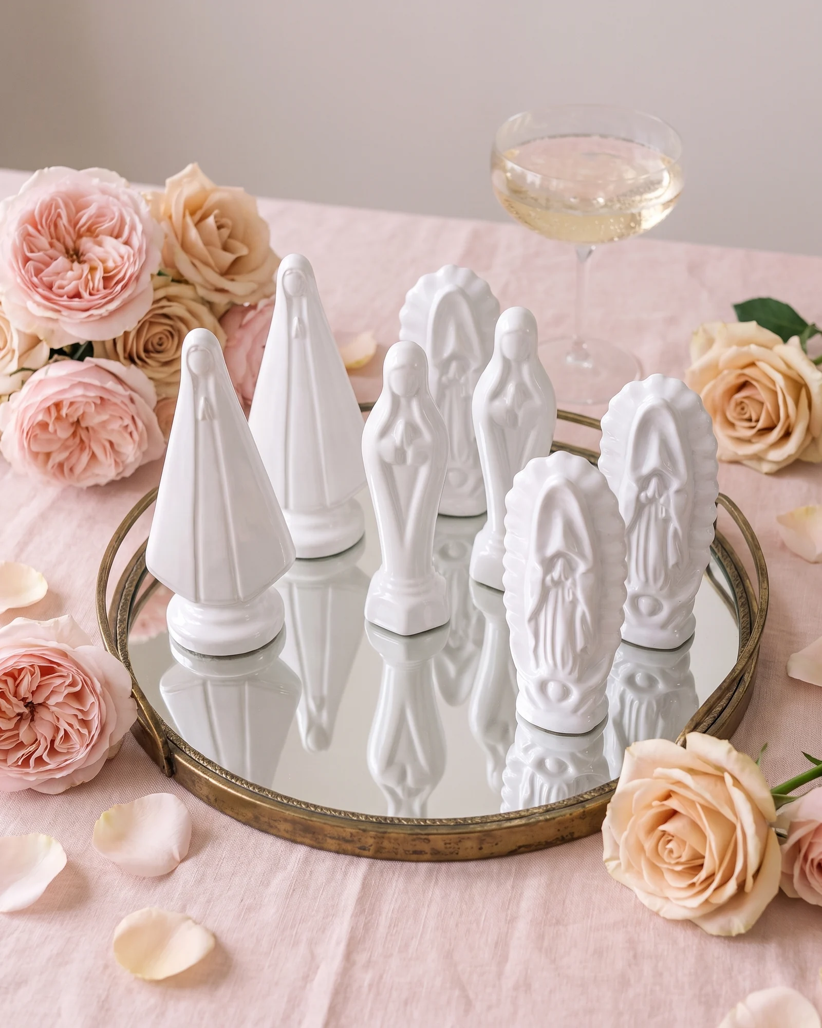 Recuerditos de cerámica blanca de la Virgen del Rosario, Madona y Silueta sobre una charola de espejo, entre rosas rosa y durazno sobre mantel rosa palo.